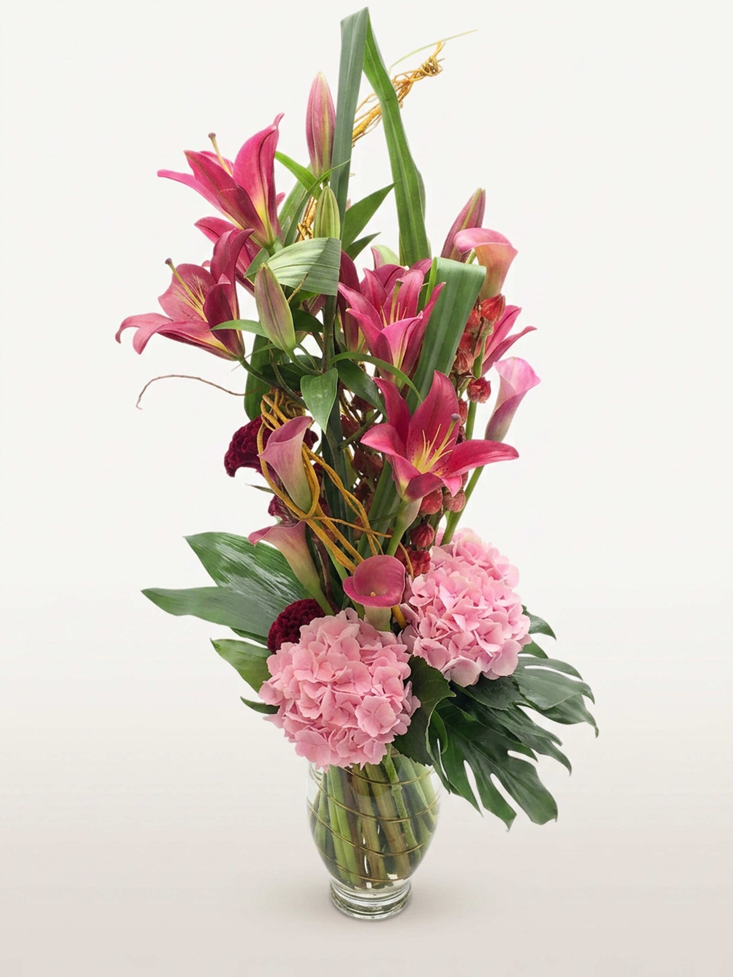 Beaux Arts : lys roses, arums, hortensias roses et feuillages verts dans un vase transparent