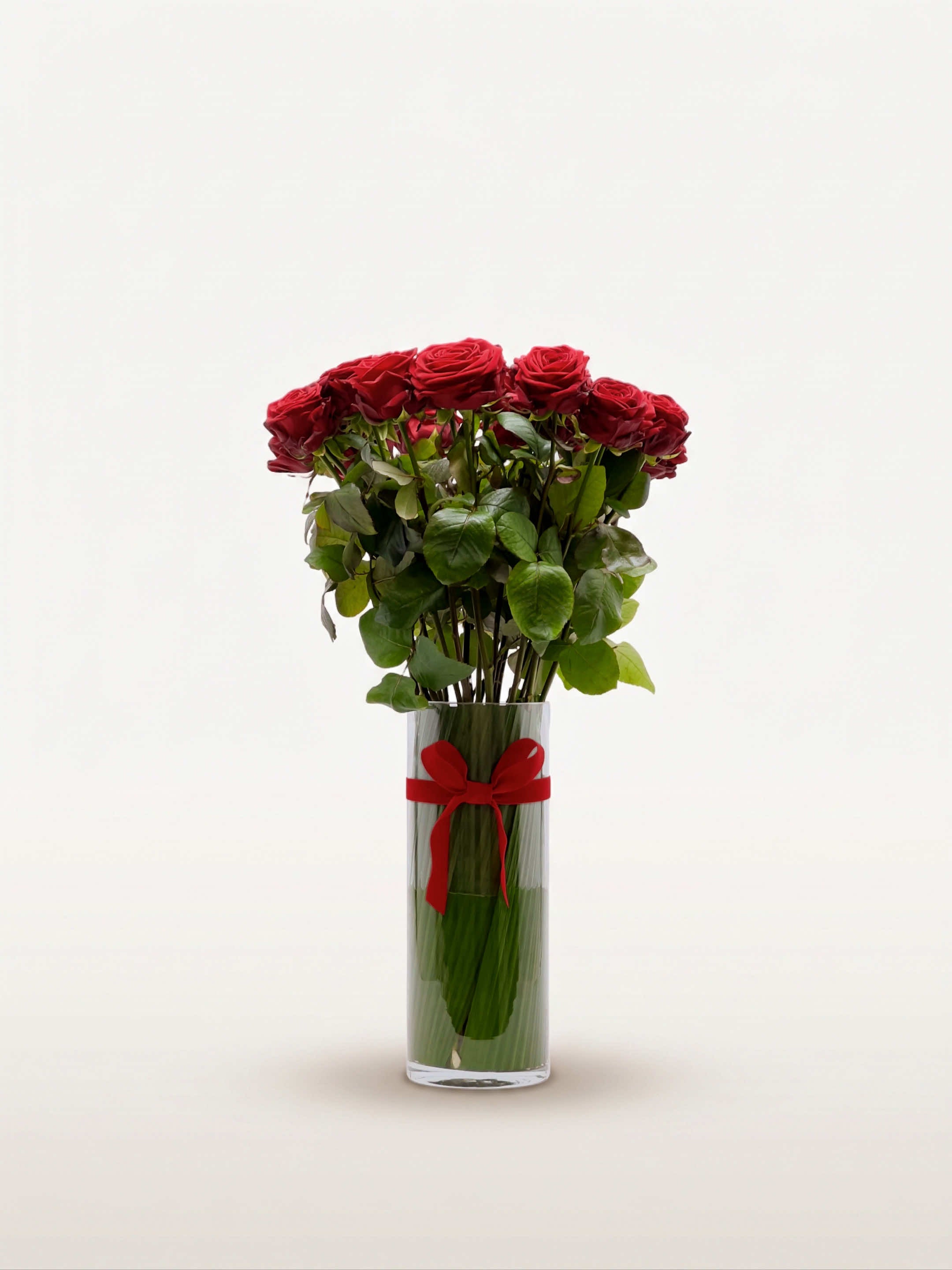 Royal N°25 rassemble une brassée de roses rouges veloutées dans un vase cylindrique en verre, noué d'un ruban rouge