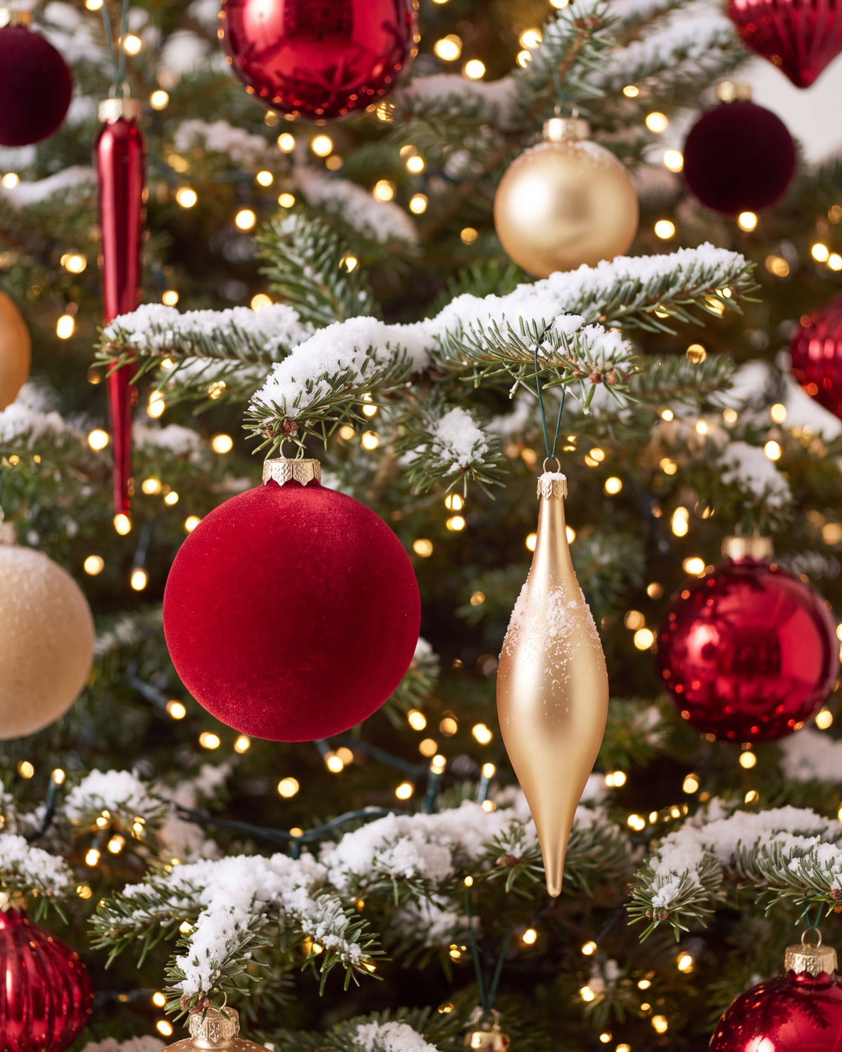 Détail du sapin de Noël Parure Opéra : boules rouges et or entre des branches enneigées et éclairées 