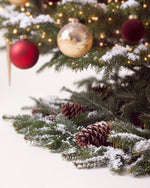 Pied du sapin de Noël Parure Opéra : branches de sapin posées au sol, pommes de pin et neige artificielle 